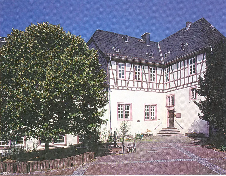 Das Foto zeigt ein großes, historisches Fachwerkgebäude mit einem hell verputzten Erdgeschoss und dunkel abgesetztem Fachwerk im oberen Bereich. Es hat ein hohes, dunkles Schieferdach mit mehreren Gauben. Vor dem Gebäude befindet sich ein gepflasterter Platz mit einer Treppe zum Haupteingang sowie einem großen Baum auf der linken Seite, der teilweise Schatten wirft. Die Szene wirkt ruhig und gepflegt, typisch für ein denkmalgeschütztes oder öffentlich genutztes Gebäude.