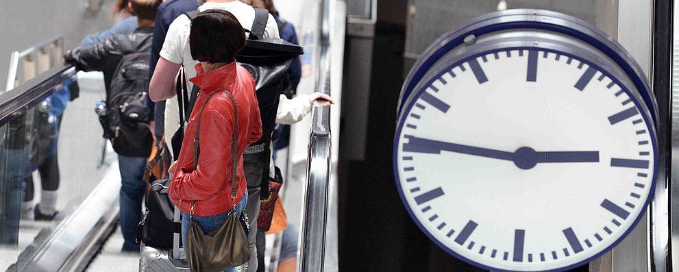 Mehrere junge Leute mit Rollkoffern stehen auf einer Rolltreppe. Rechts im Bild ist einer Uhr, sie zeigt Viertel vor drei.