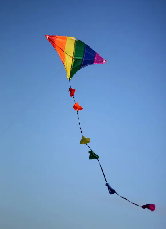 Ein Bunter, Regenbogen-farbener Flugdrache fliegt vor einem blauen Himmel.