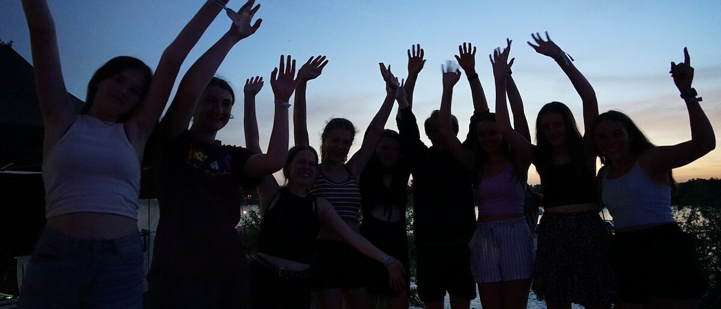 Man sieht eine Gruppe Jugendliche vor dem Abendhimmel, die Gesichter verschattet, die die Arme hoch reißen