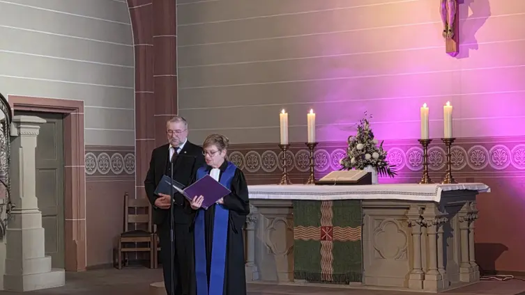 Die beiden Personen stehen vor dem Altar an einem Mikrofon. Rechts oben an der Wand eine Christusfigur am Kreuz.