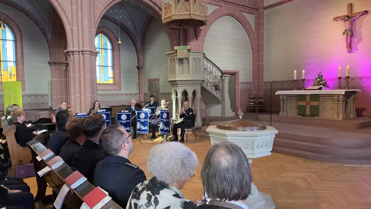 Im Altarraum der Kirche ist rechts der Altar, hinten sitzen die Musiker, vorne links Besucher in den Reihen.