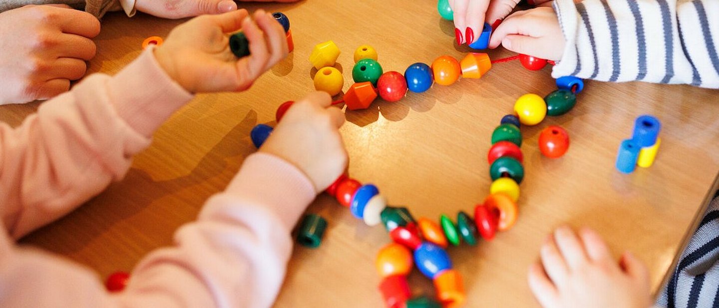 Kita - Kinder beim Spielen. An einem Tisch sitzen einige Kinder, eine Erzieherin und spielen mit einer Kette mit bunten Kugeln und anderen Formen, die auf einer Schnur aufgereiht sind.