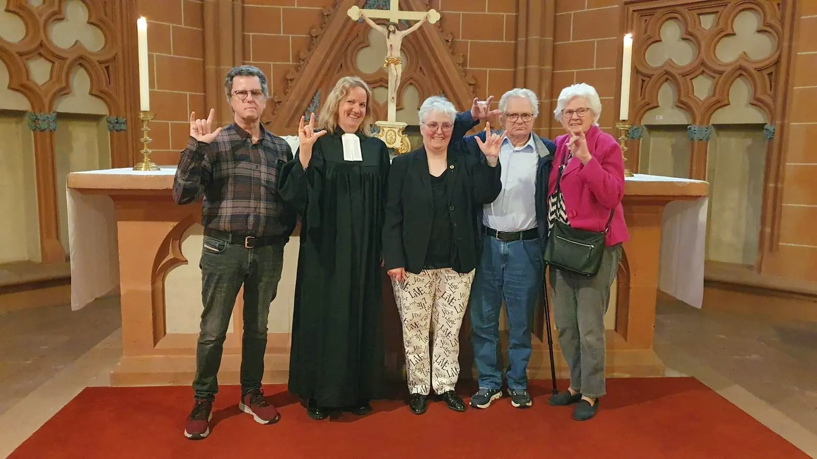 Fünf Personen stehen vor einem Altar in einer Kirche und zeigen das Gebärdenzeichen für „Ich liebe dich“.