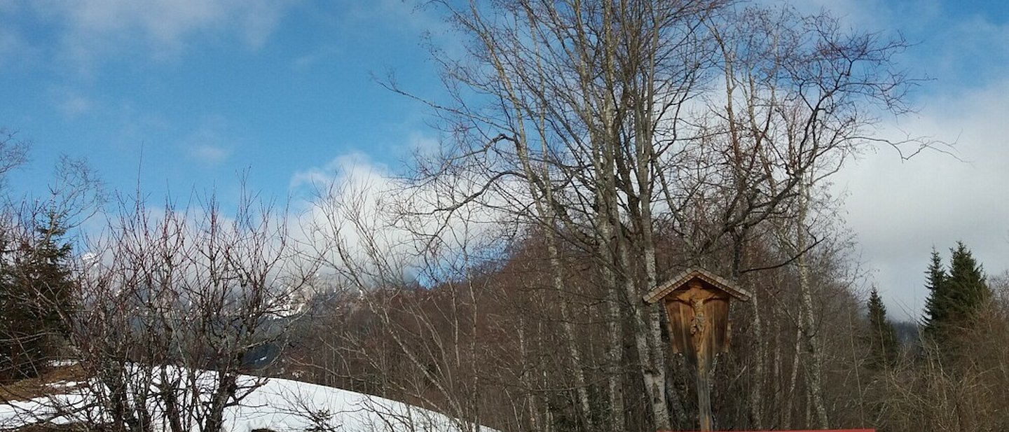 Eine Bank zum Ausruhen unter einem Marterl in verschneiter Winterlandschaft.