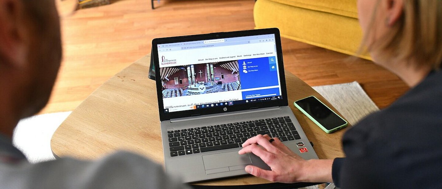 Pfarrerin Nele Schomakers und Peter Burlager - mit dem Rücken zur Kamera - schauen auf den Monitor eines Laptops. Im Hintergrund ist ein Bücherregal mit Bücher im Bild und links daneben ein Blumentopf auf einem Hocker zusehen.