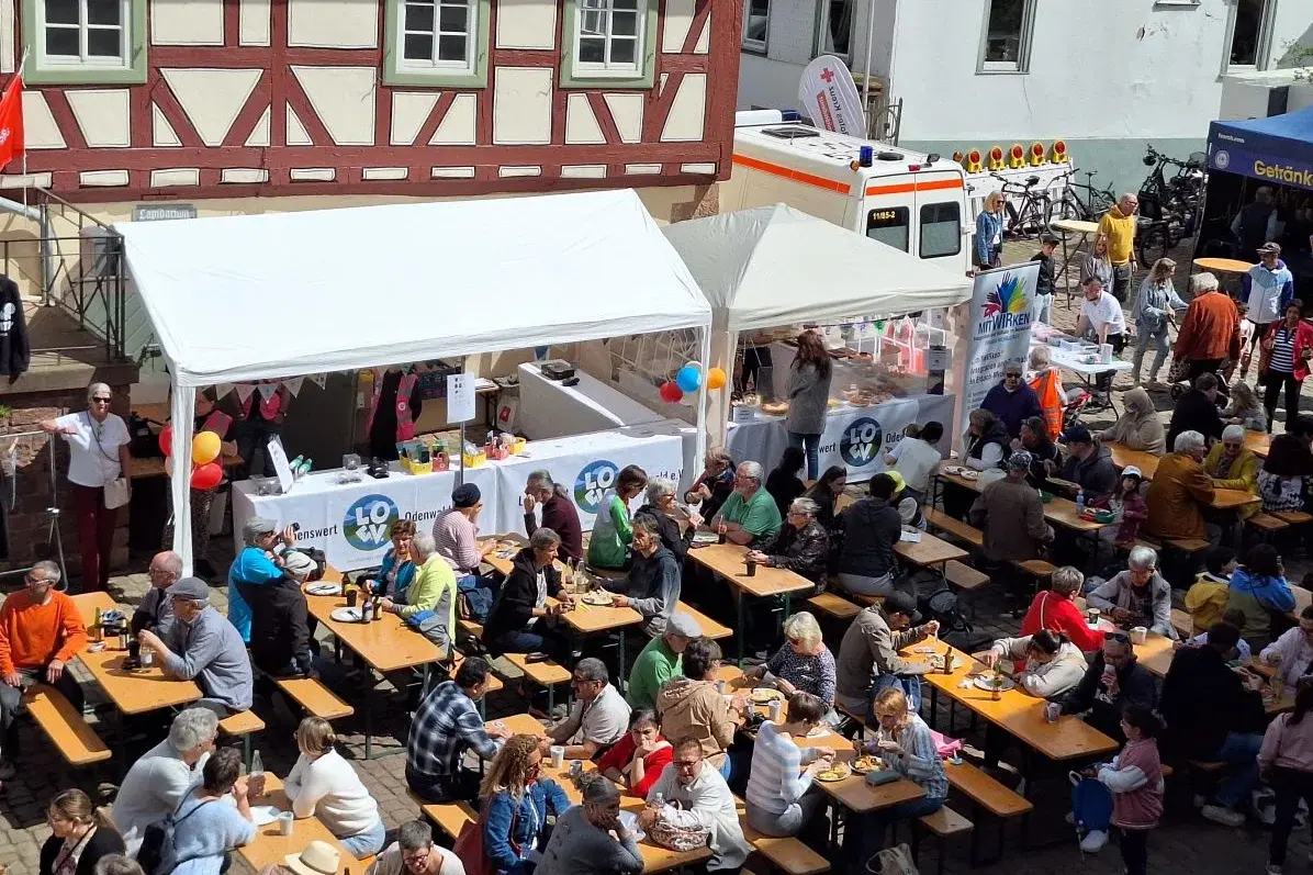 Blick von oben auf Sitzbänke mit vielen Menschen, den Kuchenstand mit weißem Zelt und Fachwerkhäusern im Hintergrund