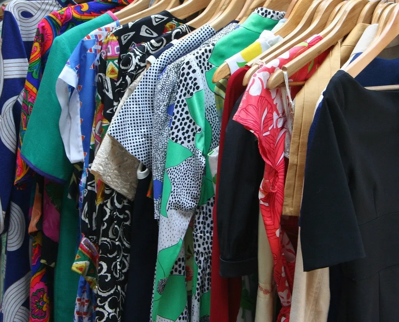 Das Bild zeigt eine Reihe von Kleidungsstücken, die an Kleiderbügeln hängen. Die Kleidungsstücke sind hauptsächlich T-Shirts und Blusen in verschiedenen Farben und Mustern.
