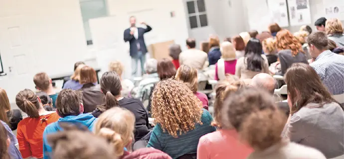 Gezeigt wird ein Hörsaal mit einer großen Gruppe sitzender Studenten, die der Vorlesung und dem Dozenten zuhören.