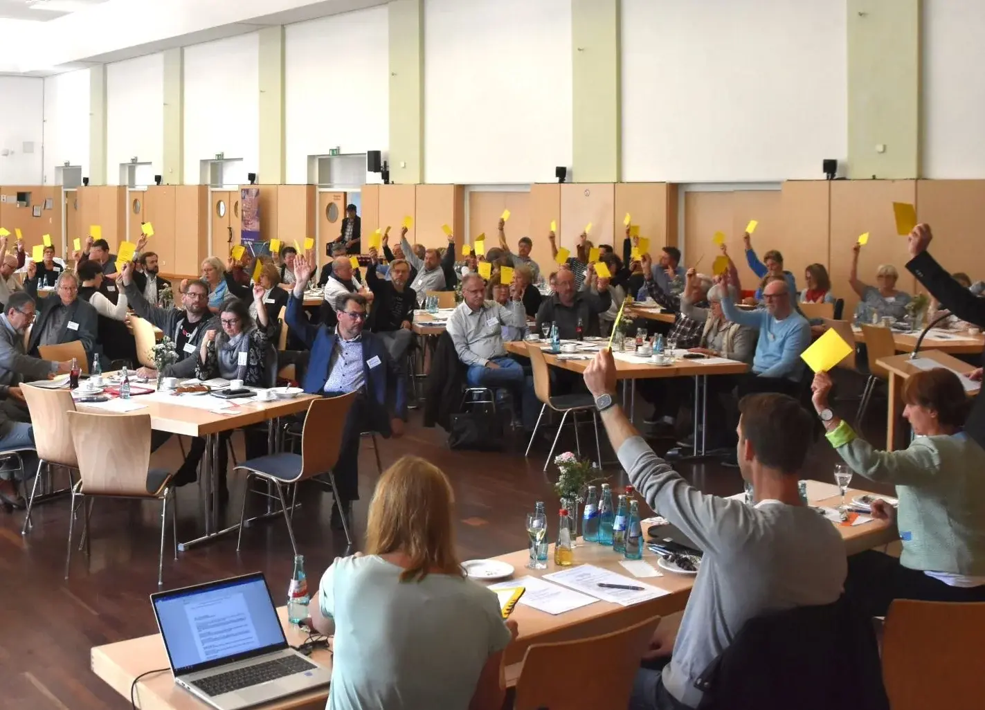 Blick in einen großen Saal, viele Menschen an Tischgruppen bei einer Abstimmung. Sie halten gelbe Stimmzettel in die Höhe.