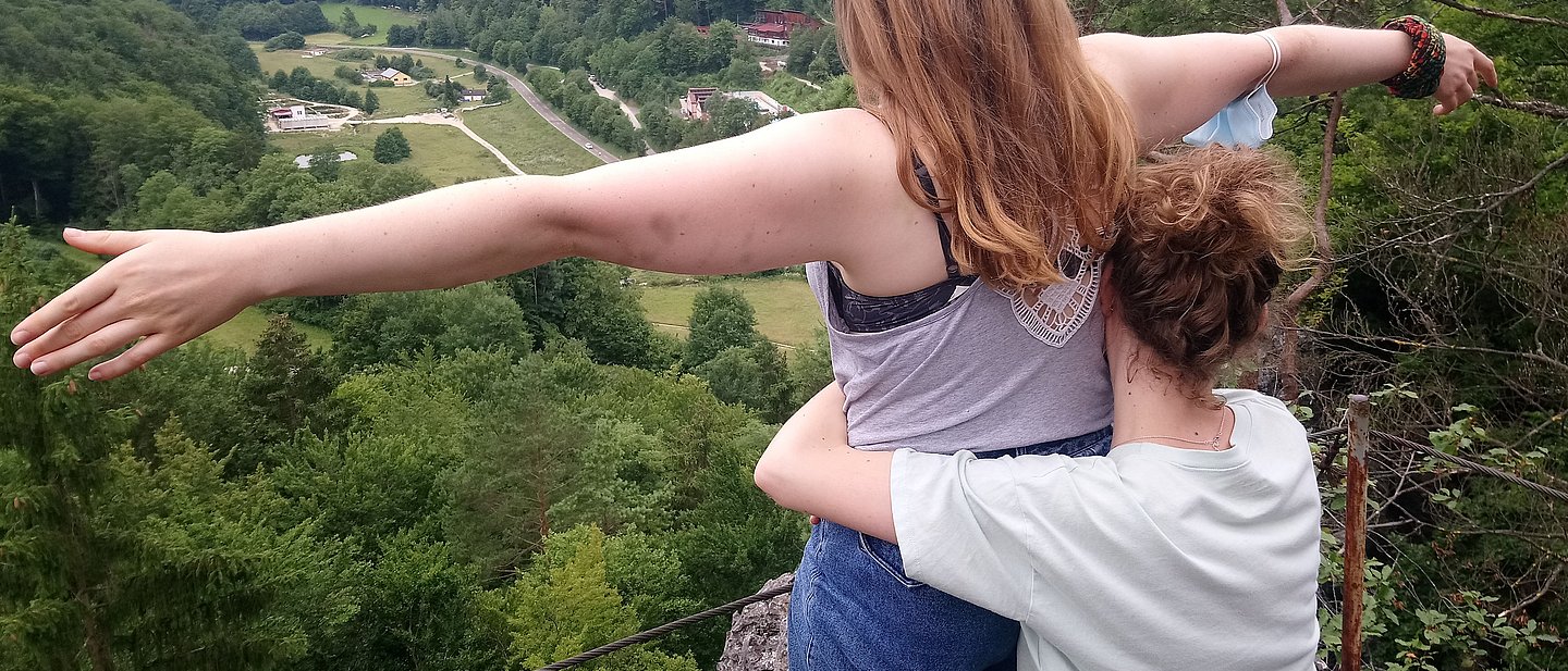 Eien Jugendliche steht am Felsrand und breitet ihre Arme glücklich aus und blickt auf ein Tal. Eine andere Jugendliche hält sie fest.