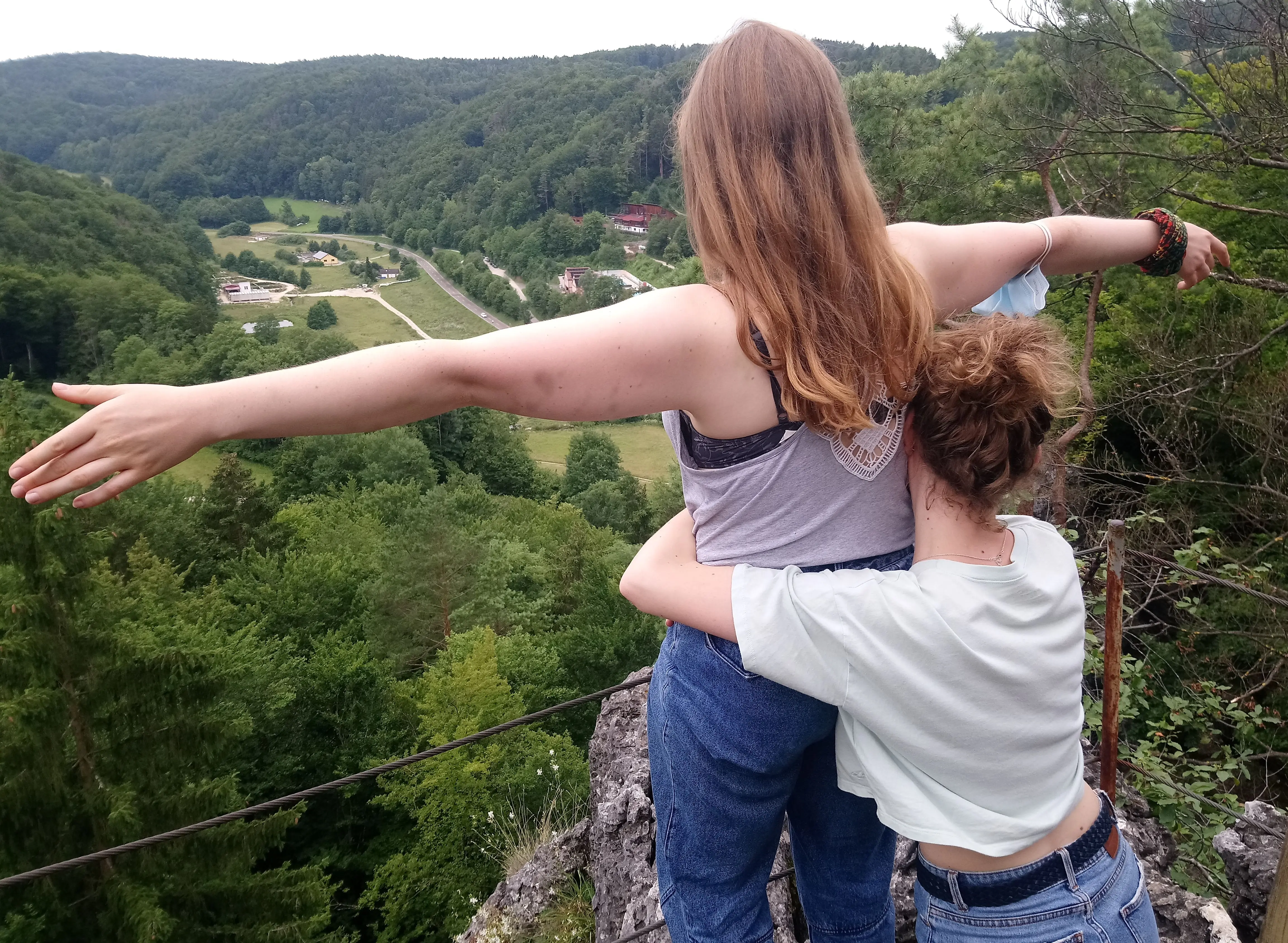 Eien Jugendliche steht am Felsrand und breitet ihre Arme glücklich aus und blickt auf ein Tal. Eine andere Jugendliche hält sie fest.