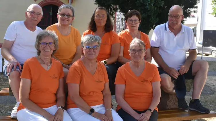 Sechs Frauen in orangefarbenen T-Shirts und zwei Männer in weißen Hemden sitzen in zwei Reihen auf einem Tisch und einer Bank vor der Niddaer Stadtkirche.