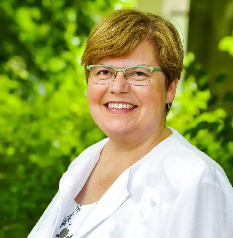 Eine Frau, Ende 50, mit braunem, kurzen Haar, grüner Brille, weißem Blazer und weißem Shirt, schaut freundlich in die Kamera,im Hintergrund Buschwerk