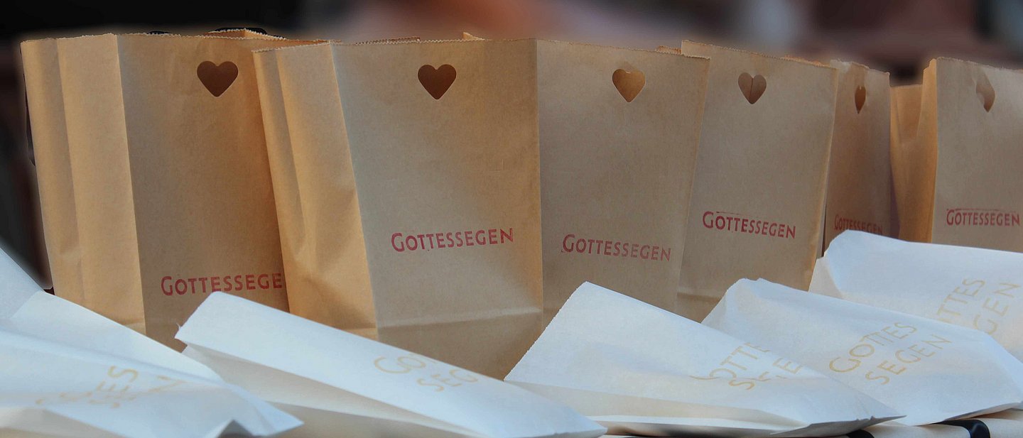 Auf dem Bild stehen braune Brottüten mit einem kleinen ausgestanzten Herz und dem Schriftzug "Gottessegen" auf einem Tisch. Davor liegen weiße Butterbrottüten