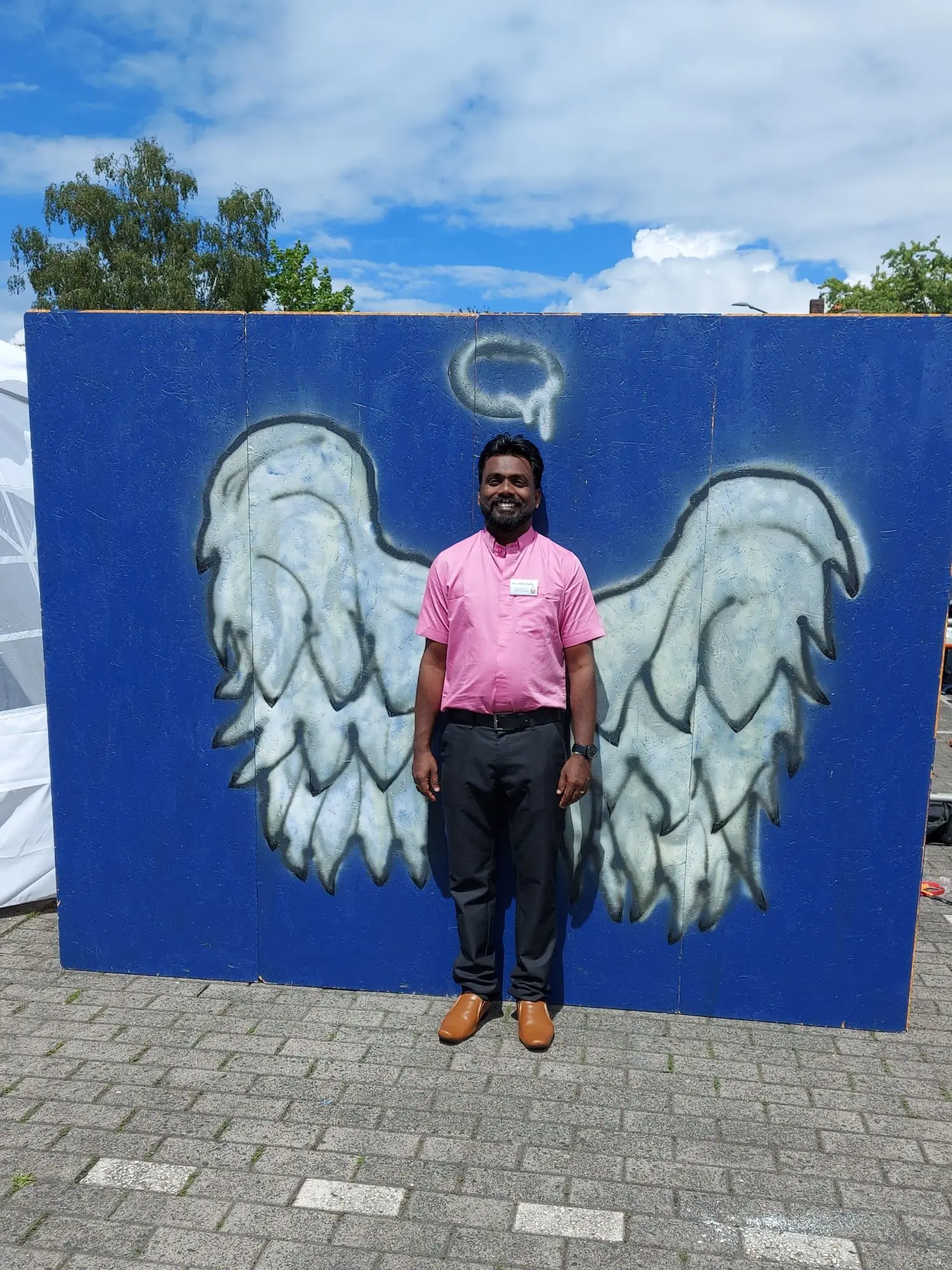 Joby Baby steht vor einer blauen Wand, auf der große Engelsflügel aufgemalt sind.