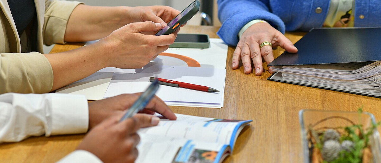 Sabine Schlösser, Luise Pahl und Benjamin Colin Köpper sitzen an einem Tisch während eder Arbeit. Man sieht nur Hände und Arme, keine Gesichter. Auf dem Tisch liegt ein Ordner und steht eine Dekoschale. In ihren Händen halten die Personen Handys.