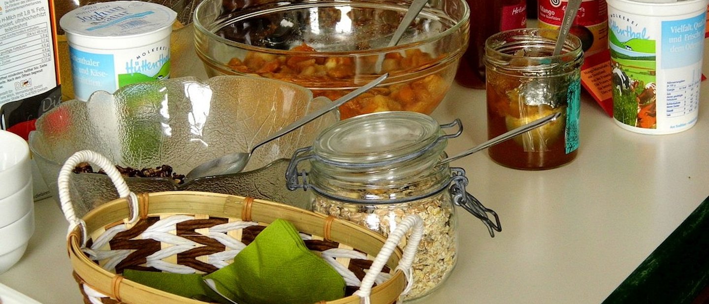 Zu sehen ist ein Buffet-Tisch, auf welchem sich eine Auswahl an Frühstücksessen befindet: Müsli, Marmelade, ein Brotkörbchen, Obstsalat, Kuchen, und Jogurt.