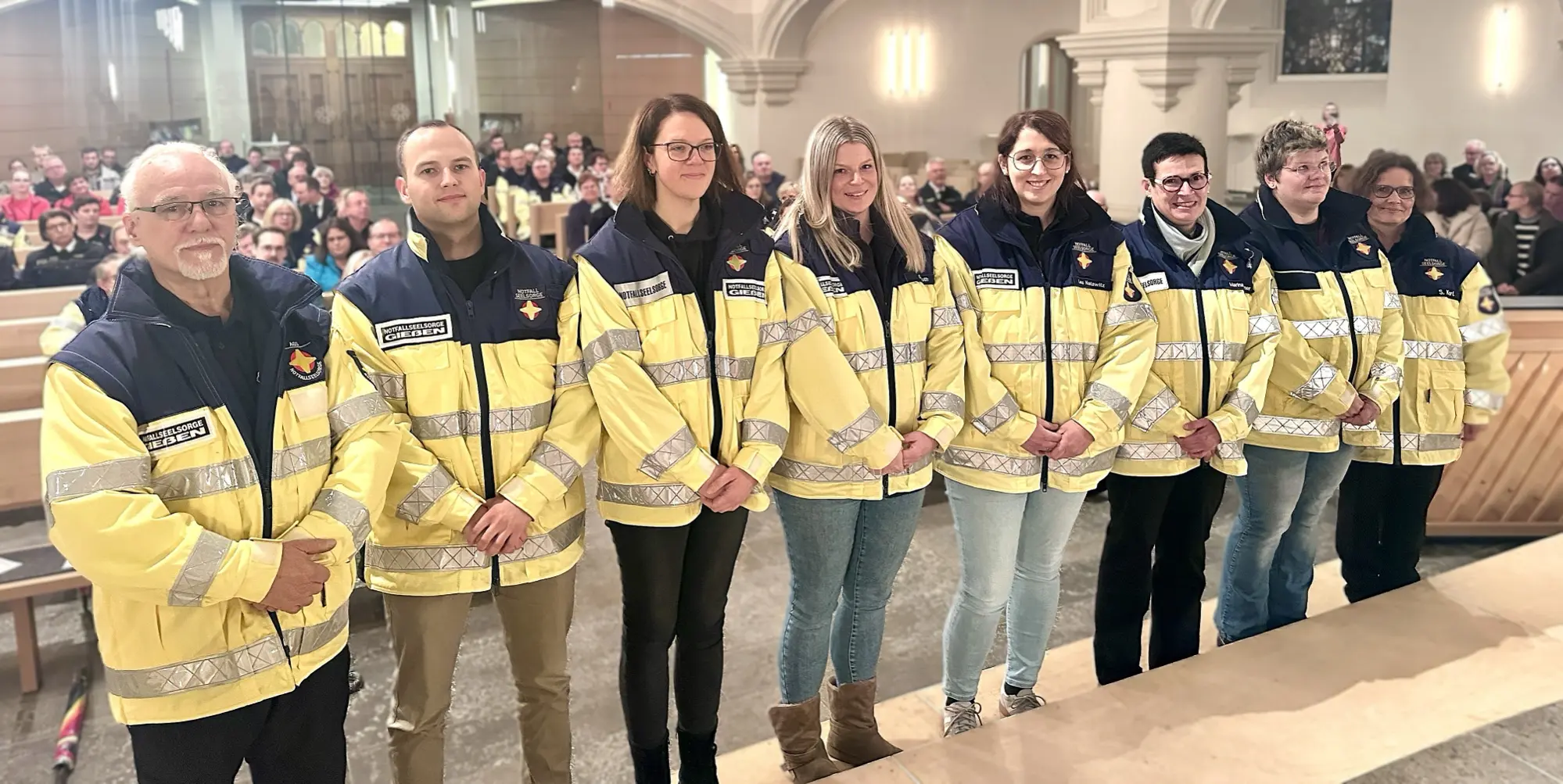 In einer Kirche steht eine Gruppe von acht Einsatzkräften der Notfallseelsorge nebeneinander. Sie tragen gelbe Einsatzjacken mit reflektierenden Streifen. Im Hintergrund sitzen zahlreiche Besucherinnen und Besucher in den Kirchenbänken. Die Szene wirkt feierlich und ruhig, passend zu einem Gottesdienst.