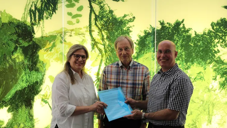 Die drei Personen stehen vor dem gelb-grünen Kunstwerk, das Naturmotive zeigt.