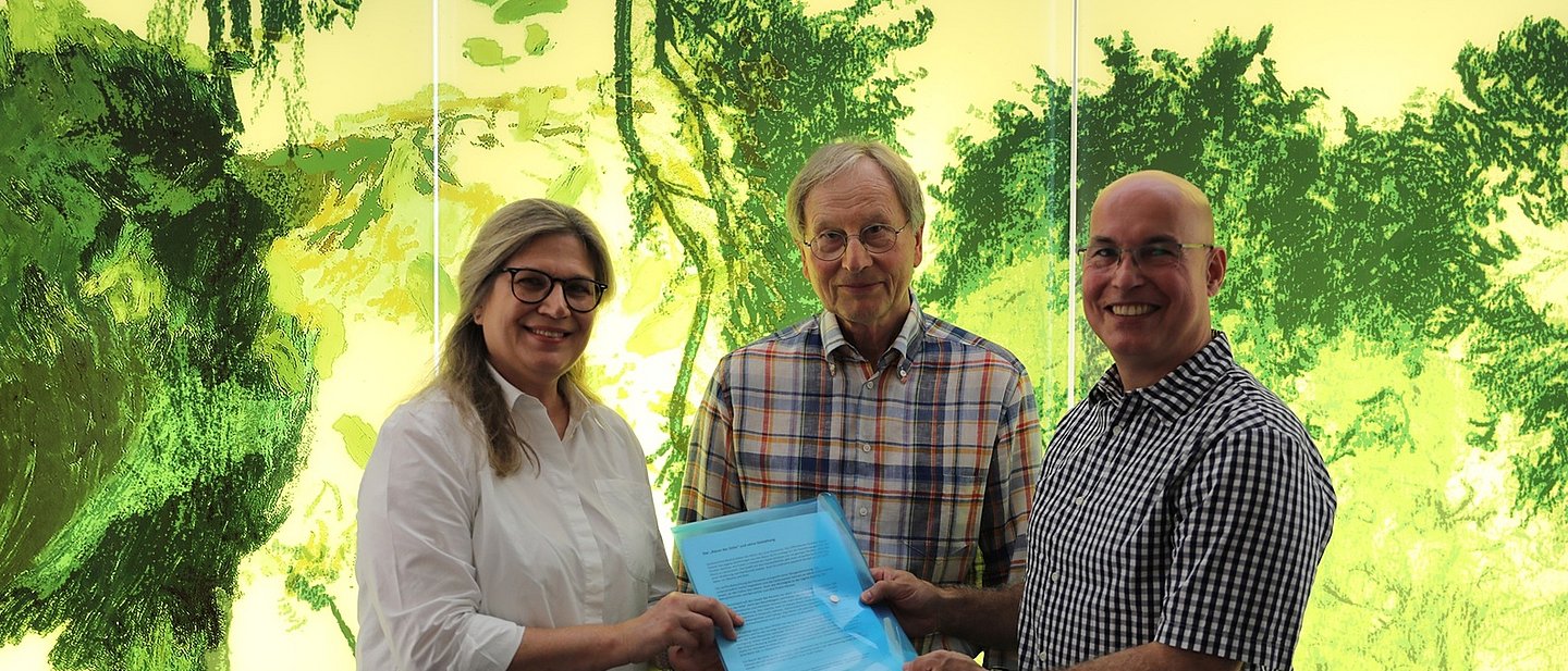 Die drei Personen stehen vor dem gelb-grünen Kunstwerk, das Naturmotive zeigt.