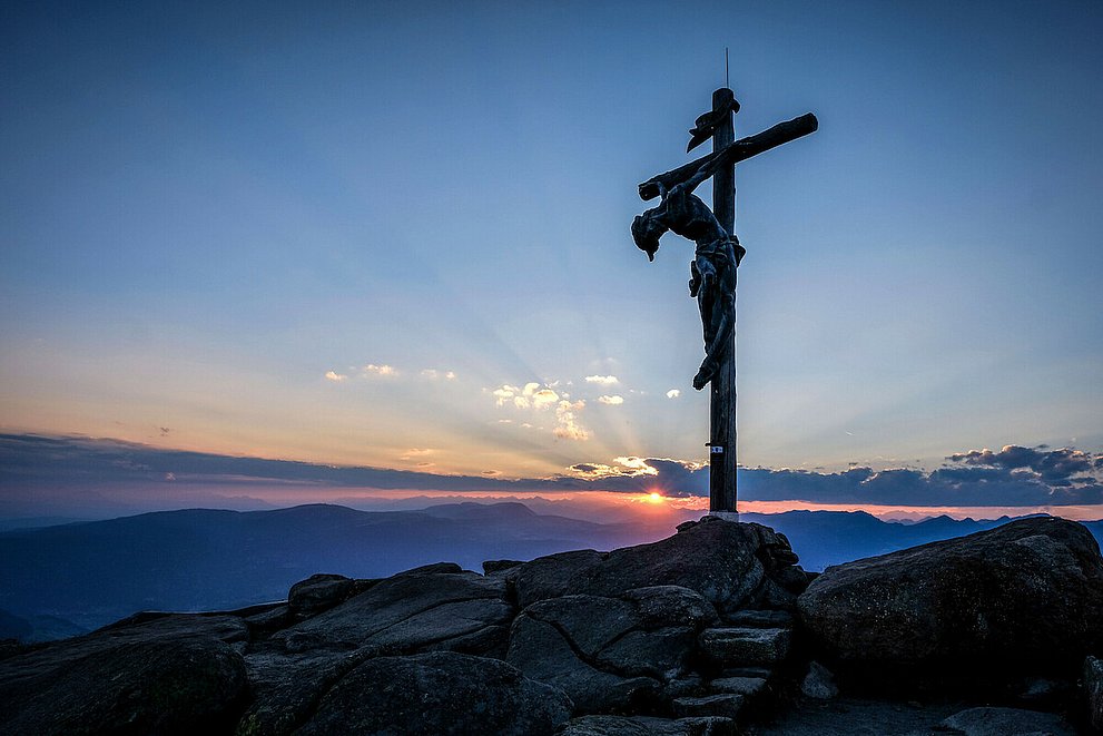 Man sieht einen Berggipfel mit dem dem überlebensgroßen Christus am Kreuz vor einem Abendhimmel mit Sonnenuntergang