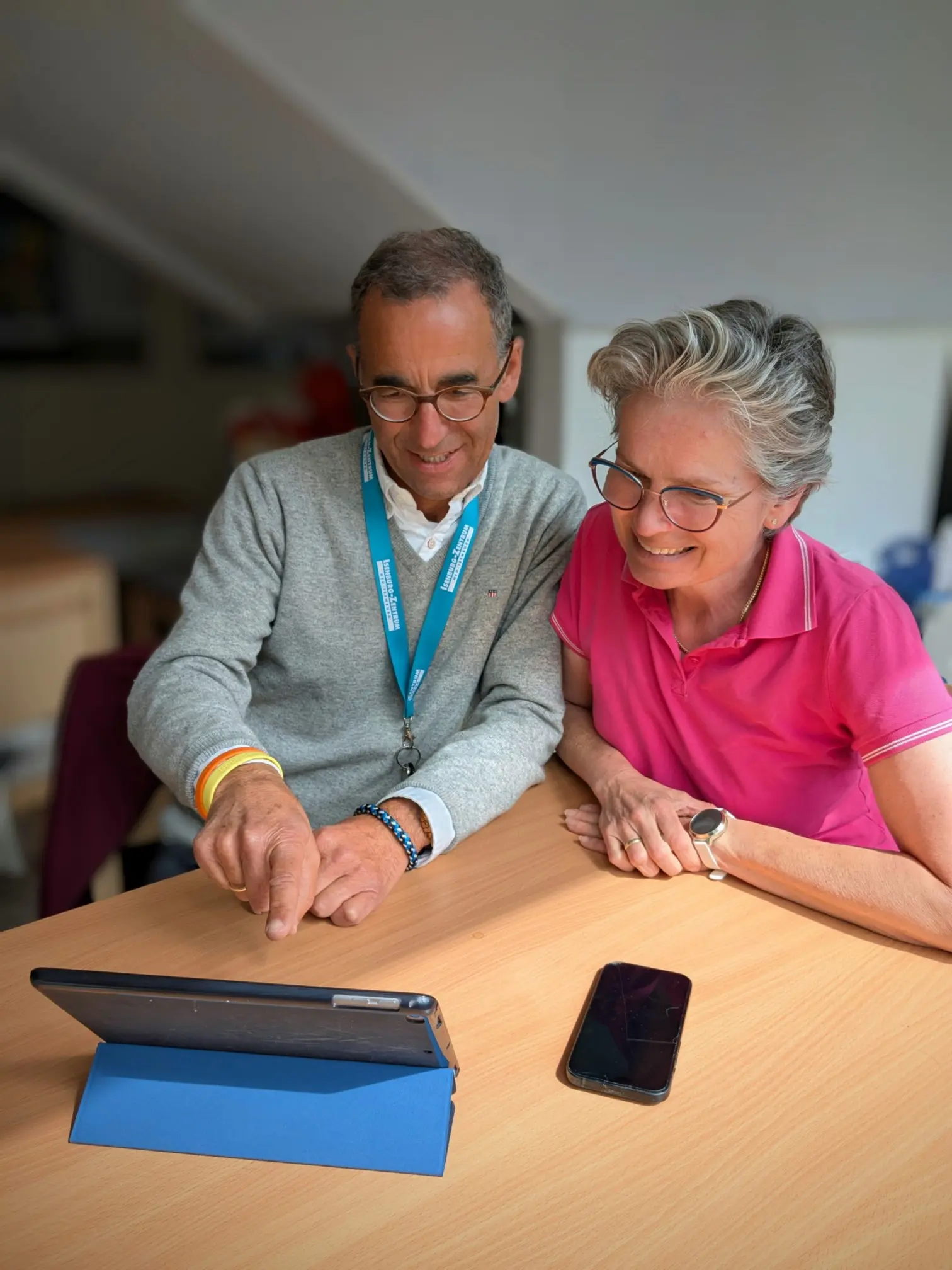 Das Bild zeigt einen älteren Mann und eine ältere Frau, die gemeinsam auf einen Tablet-Computer schauen. Der Mann trägt eine Brille, ein graues Sweatshirt über einem weißen Hemd mit einer blauen Halskette und ein gelbes Armband. Er deutet mit dem Finger auf den Bildschirm des Tablets. Die Frau, die ebenfalls eine Brille trägt, ist in ein pinkfarbenes Poloshirt gekleidet und schaut interessiert zu. Auf dem Tisch vor ihnen befinden sich das Tablet in einem blauen Ständer und ein Smartphone.