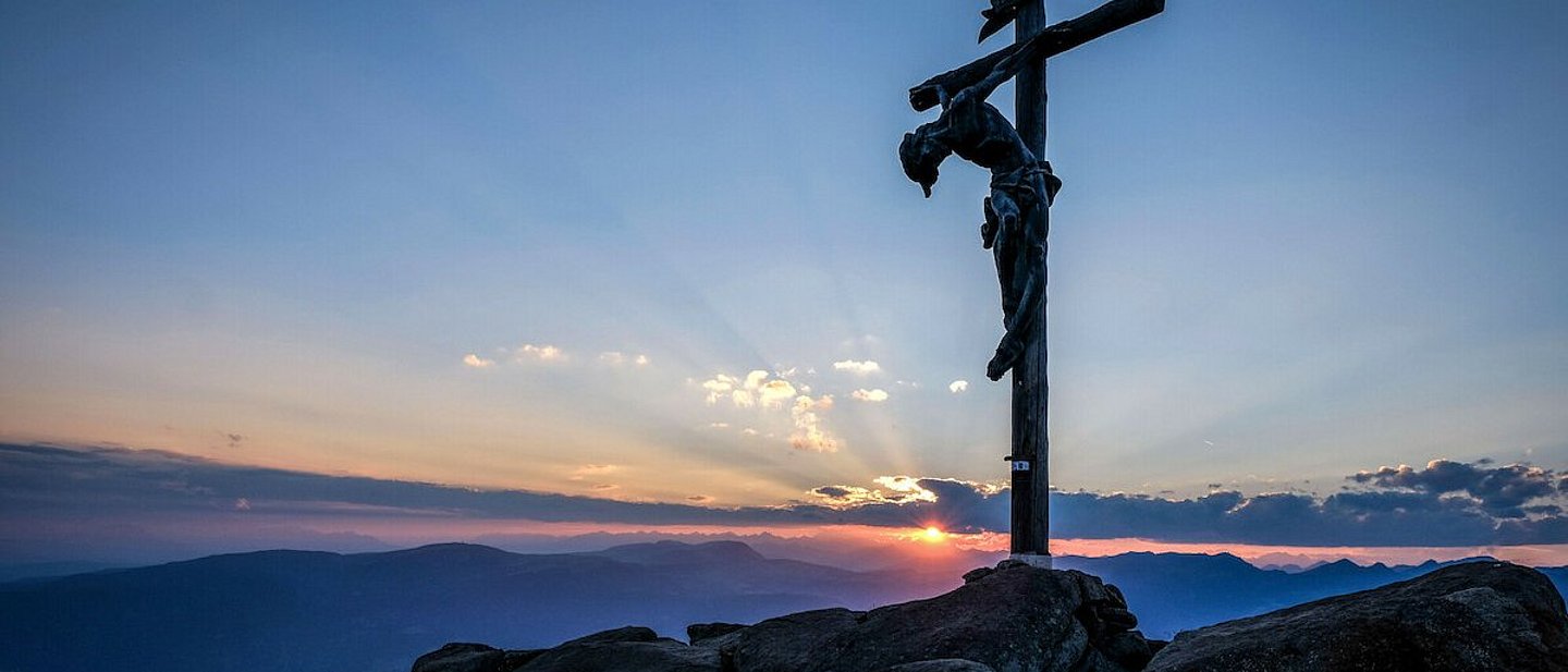 Man sieht einen Berggipfel mit dem dem überlebensgroßen Christus am Kreuz vor einem Abendhimmel mit Sonnenuntergang