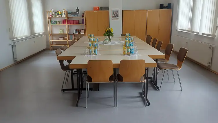Ein heller Raum. In der Mitte steht ein großer Tisch, um den Tisch herum stehen Stühle