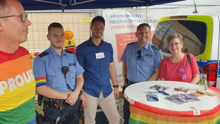 Mehrere Personen stehen um einen Bistrotisch, links der Minister, rechts die Pfarrerin, dazwischen 3 Polizisten.