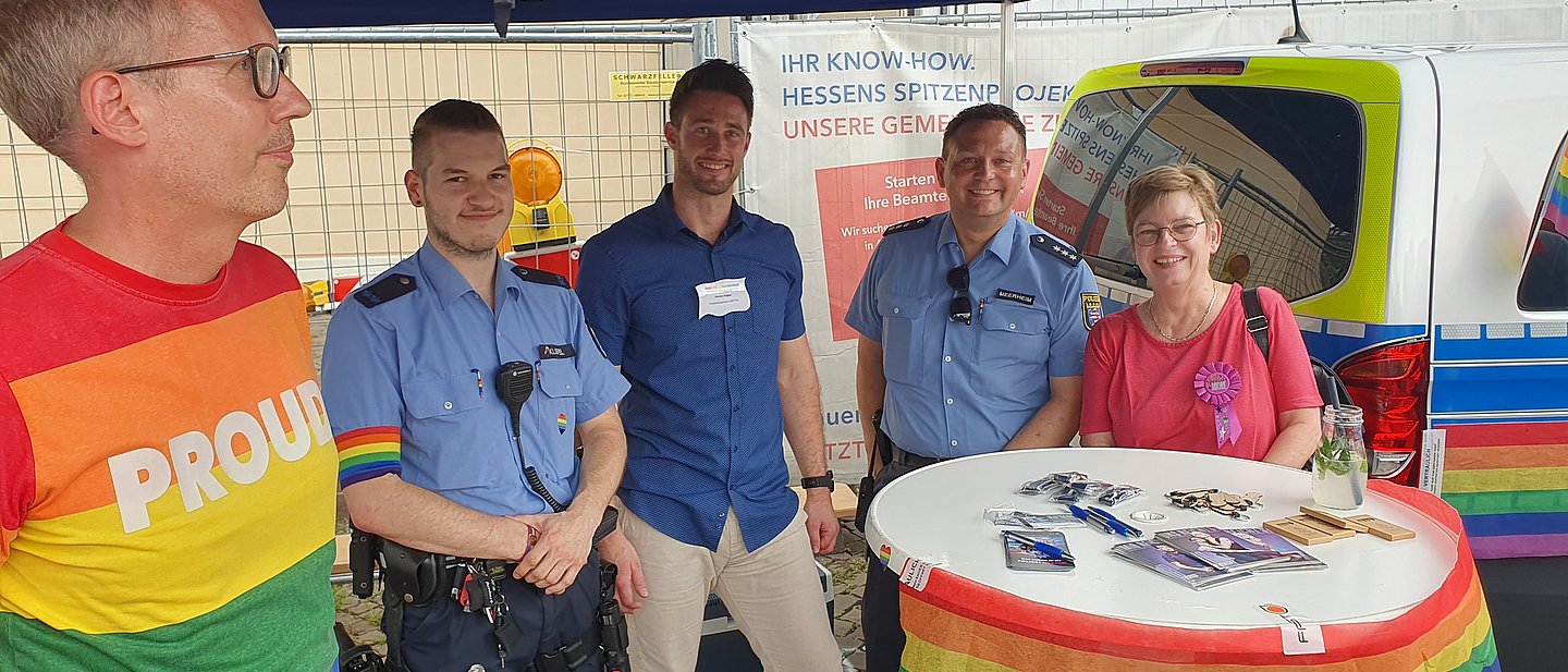 Mehrere Personen stehen um einen Bistrotisch, links der Minister, rechts die Pfarrerin, dazwischen 3 Polizisten.