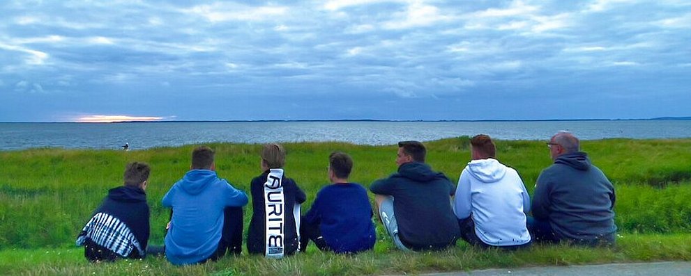 Man sieht eine Gruppe Jugendlicher am Strand sitzen die einem Kiter bei Ihrem Sport zusehen. Im Hintergrund sieht man den mit Gras bewachsenen Strand mit dem Meer dahinter. Der Drachen des Kiters fliegt in dem bewölktem Himmel auf der linken Seite des Bildes.