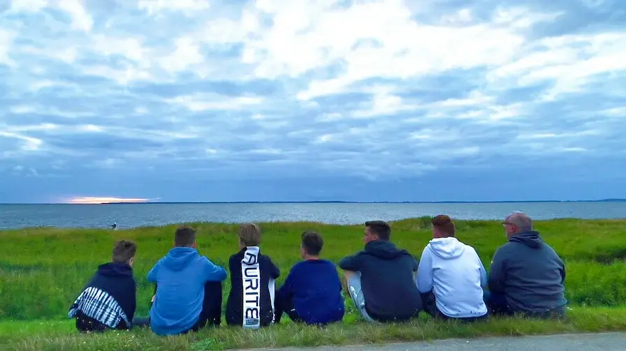 Man sieht eine Gruppe Jugendlicher am Strand sitzen die einem Kiter bei Ihrem Sport zusehen. Im Hintergrund sieht man den mit Gras bewachsenen Strand mit dem Meer dahinter. Der Drachen des Kiters fliegt in dem bewölktem Himmel auf der linken Seite des Bildes.
