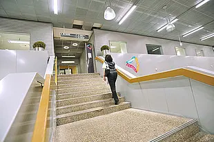 Eine Person geht eine breite Treppe in einem modernen Flughafengebäude hinauf. Helle Beleuchtung, Geländer und ein Hinweisschild mit Herzsymbol schaffen eine freundliche und offene Atmosphäre.