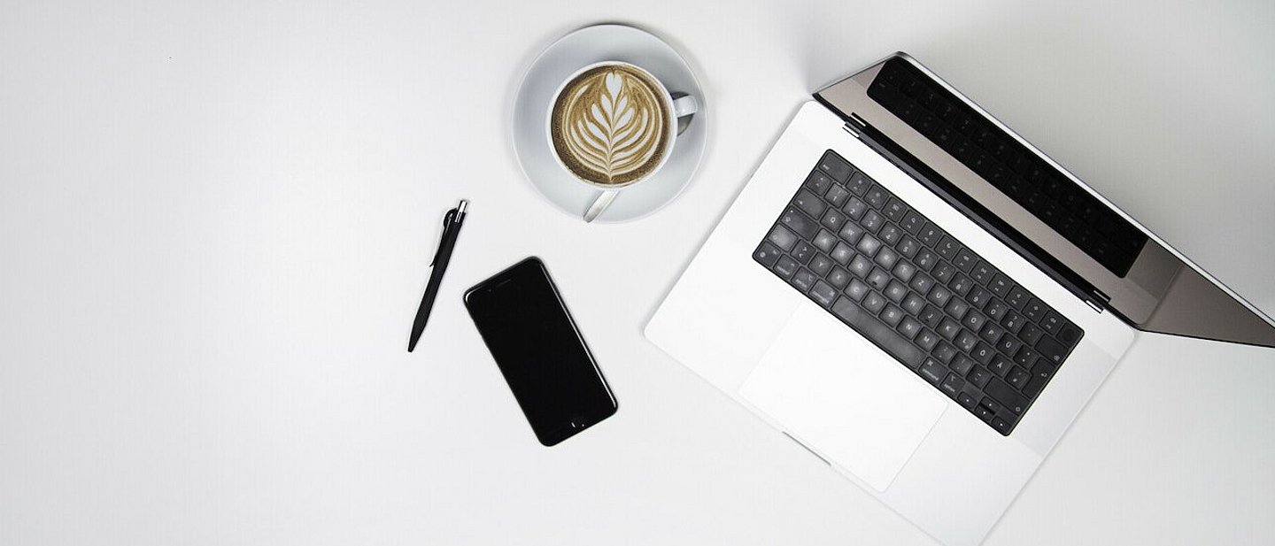 Arbeitstisch von oben mit Laptop, Kaffeetasse, Smartphone und Stift.