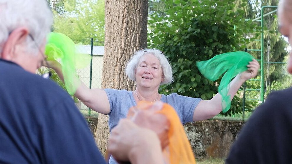 Dorothee Ewald steht strahlend vor Senior*innen und hält zwei grüne Tücher in die Höhe.