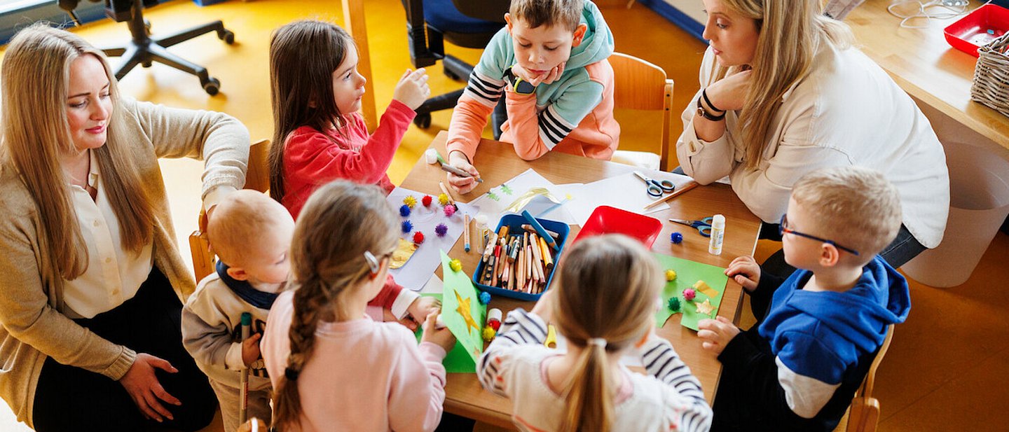 Ev. Kindertagesstätte "Kinder Garten Eden" Gemünden. Blick auf einen Tisch mit spielenden Kindern. Es sitzt noch eine Erzieherin mit am Tisch. Eine weitere Erzieherin kniet auf dem Teppich.