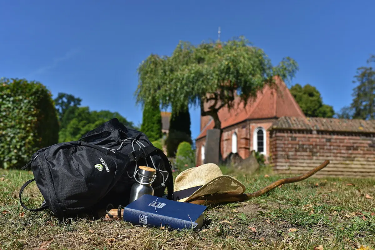Auf dem Gras vor einer Kirche liegt alles, was der Pilgerer für einen Ausflug braucht: Rucksack, Hut, Stock, Getränkeflasche und die Bibel.