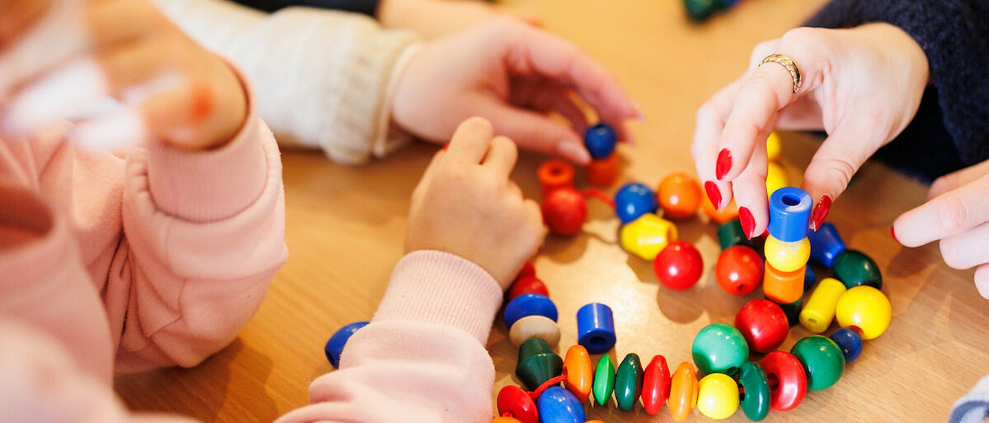 Kita - Kinder beim Spielen. An einem Tisch sitzen einige Kinder, eine Erzieherin und spielen mit einer Kette mit bunten Kugeln und anderen Formen, die auf einer Schnur aufgereiht sind.