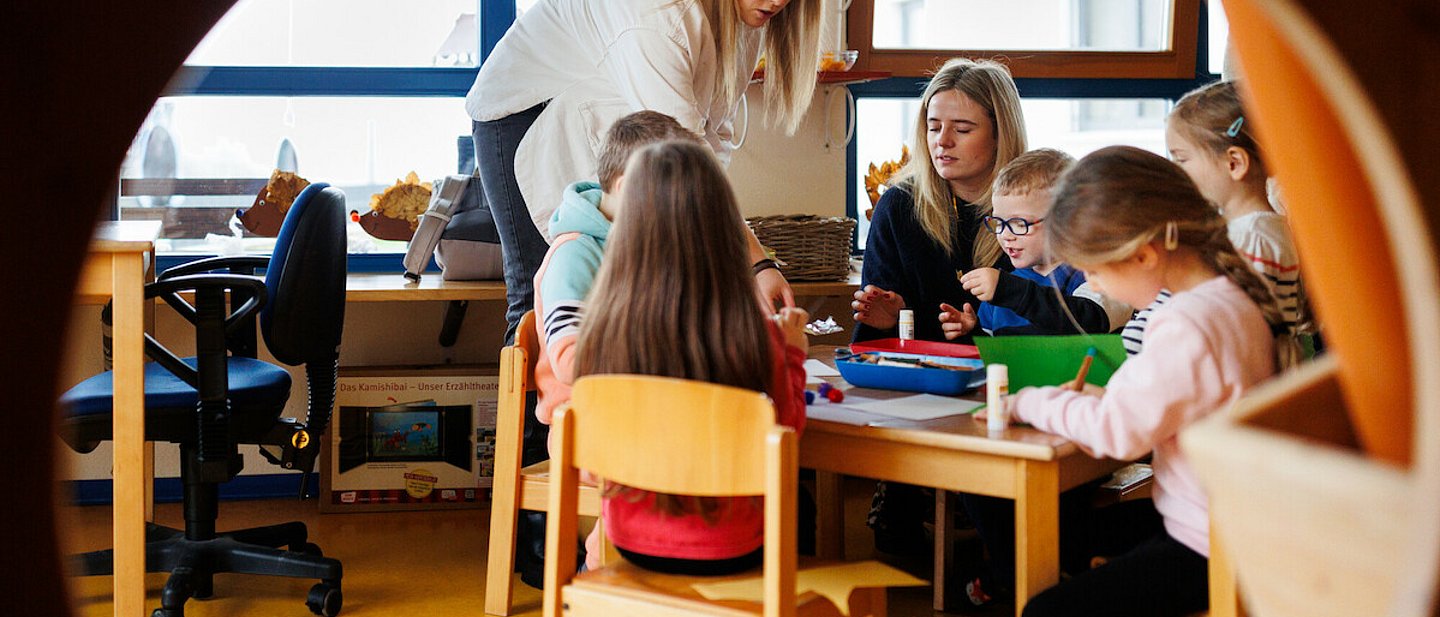 Ev. Kindertagesstätte "Kinder Garten Eden" Gemünden. Blick durch einen Kreis auf einen Tisch mit spielenden Kindern. Es sitzt noch eine Erzieherin mit am Tisch. Eine weitere Erzieherin steht am Tisch. 