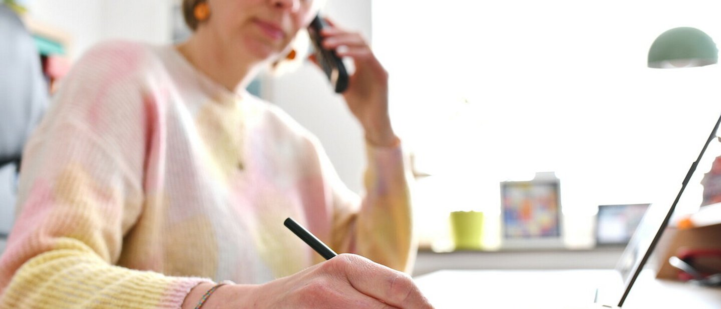 Pfarrerin Nele Schomakers in einem Büro beim Telefonieren. In der linken Hand das Telefon, in der rechten Hand ein Kugelschreiber um Notizen zu machen. Im Hintergrund ein Bücherregal - auf dem Fensterbrett ein Kreuz und ein Blumentopf.