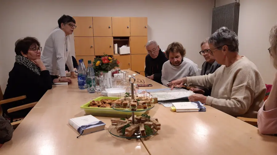 An einem Tisch sitzen mehrere Personen. Eine Frau zeigt den anderen etwas auf einer Karte. In der Mitte stehen verschiedene Leckereien.