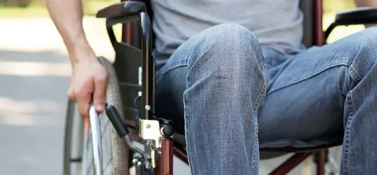 Ein Mann im Rollstuhl, von der Seite aufgenommen. Er trägt Jeans und ein graues T-Shirt und greift mit der Hand an das Rad seines Rollstuhls.