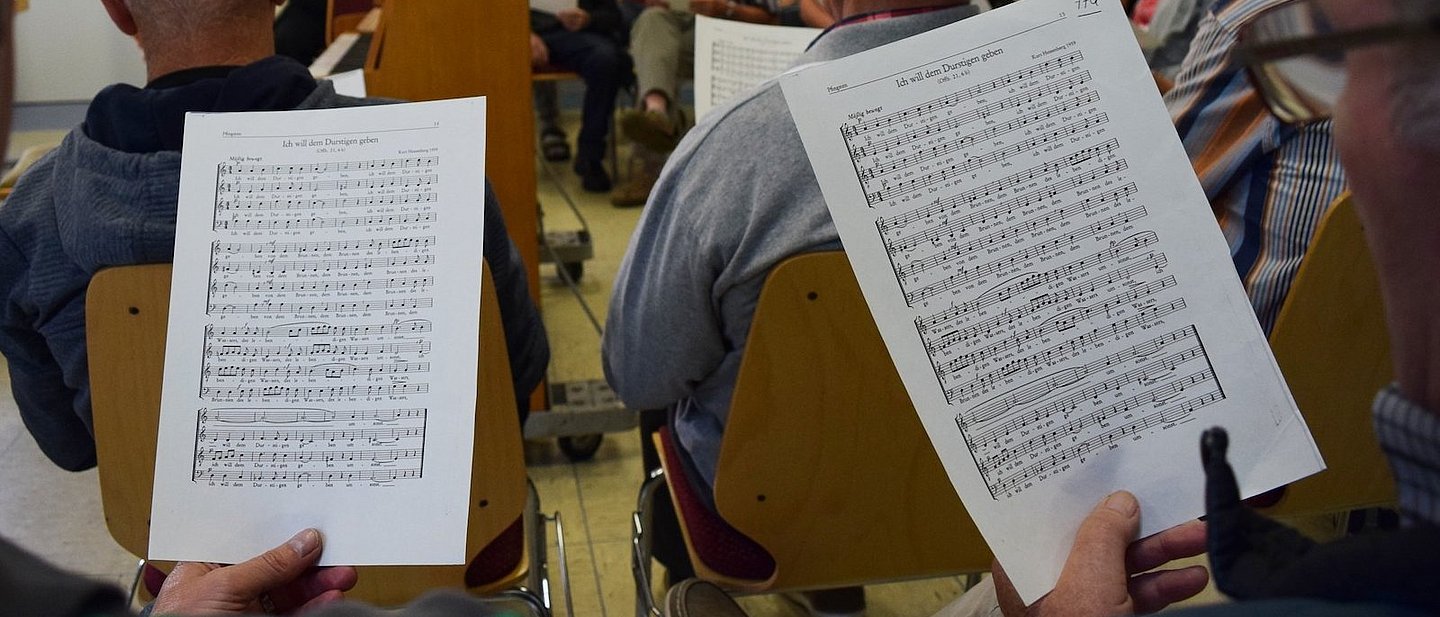 Probe in einem Kirchenchor: zwei Männer mit Noten im Vordergrund, dahinter die Dirigentin und andere Chormitglieder.