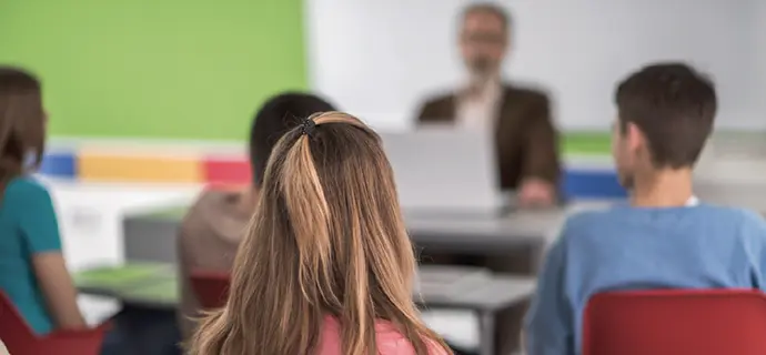 Blick von der letzten Schulbank, an Schülerinnen und Schüler vorbei in Richtung Tafel und Lehrer, der an einem Laptop sitzt.