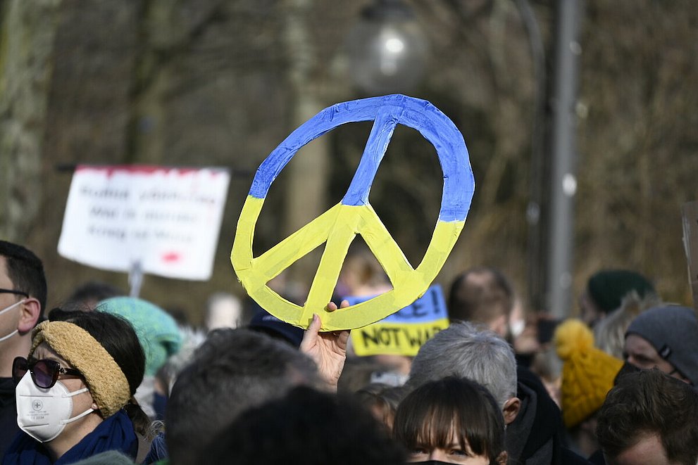 Ein Demonstrationszug gegen den Krieg in Ukraine. Ein Demonstrierender hält ein Schild mit einem Friedenszeichen hoch, das in ukrainischen Farben angemalt ist.