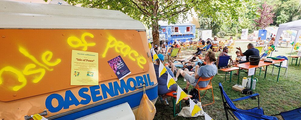 Man sieht im Vordergrund das Oasenmobil und im Hintergrund Jugendliche auf Bänken und Stühlen