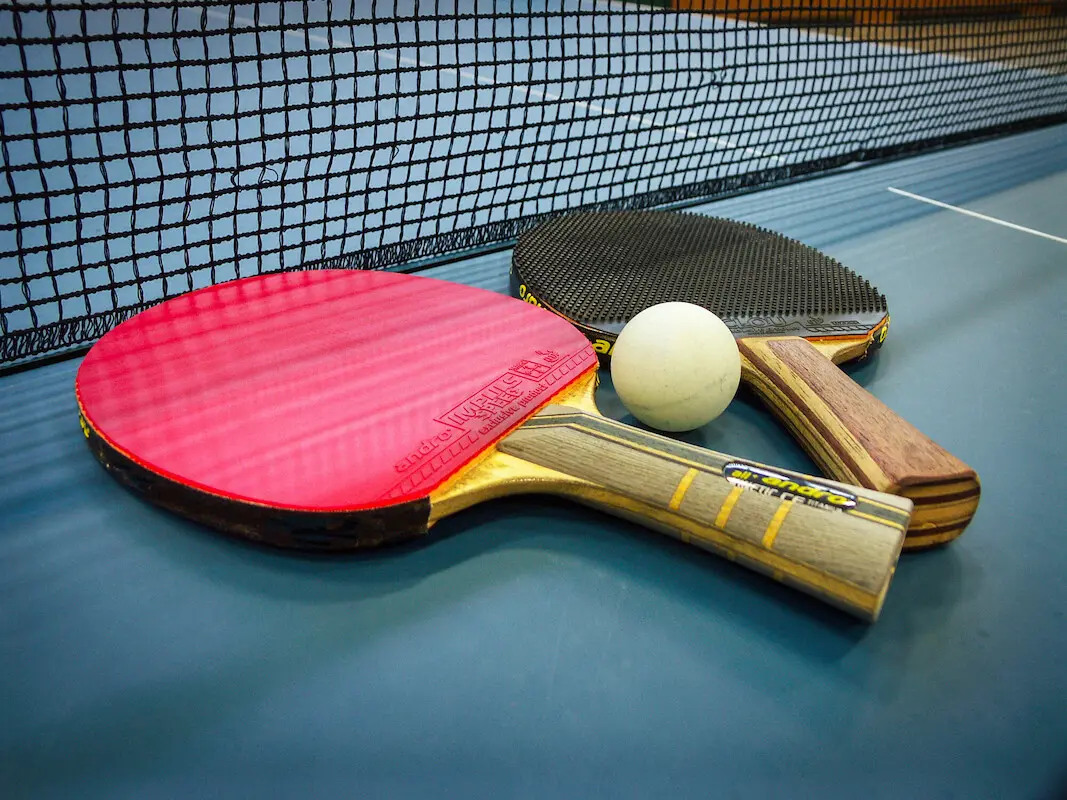 Zwei Tischtennisschläger. jeweils mit einem roten und einen weißen Belag und ein Ball liegen vor dem Netz auf der grünen Tischtennisplatte