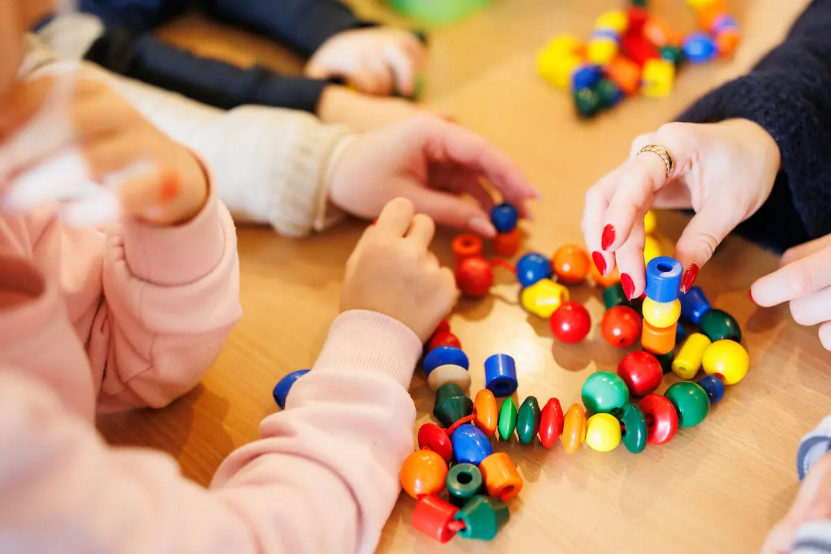 Kita - Kinder beim Spielen. An einem Tisch sitzen einige Kinder, eine Erzieherin und spielen mit einer Kette mit bunten Kugeln und anderen Formen, die auf einer Schnur aufgereiht sind.