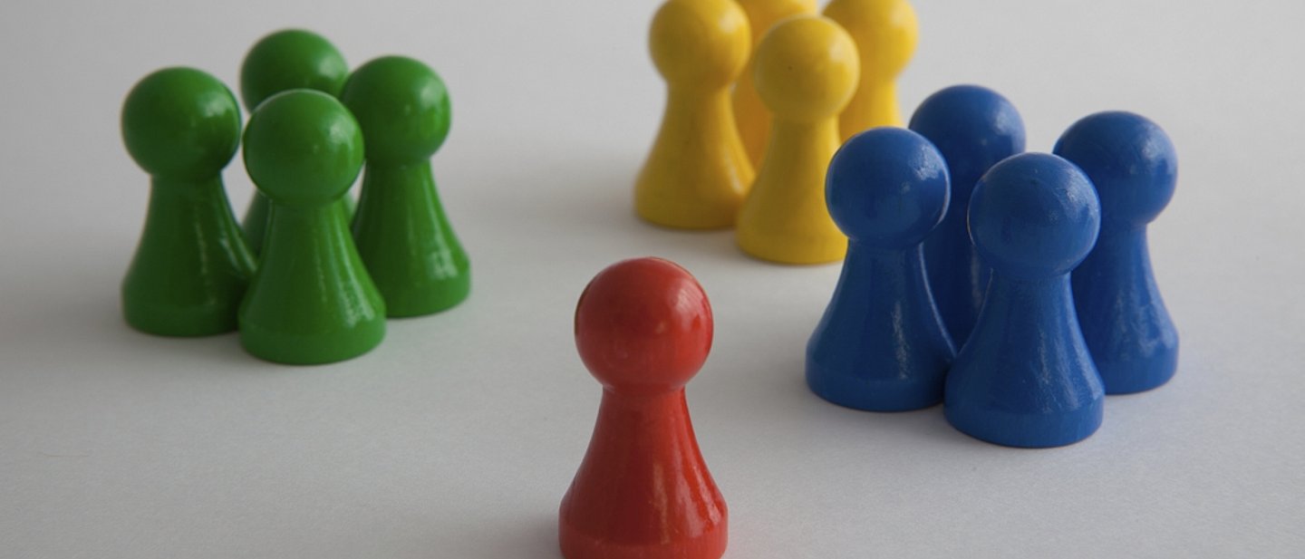 Eine rote Spielfigur steht vor drei Gruppen mit jeweils vier grünen, gelben und blauen Spielfiguren